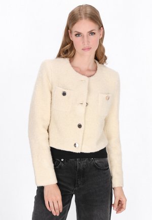 Cream-colored cropped jacket with a soft, textured fabric, round neckline, two front pockets, and silver-tone buttons. Paired with black pants.