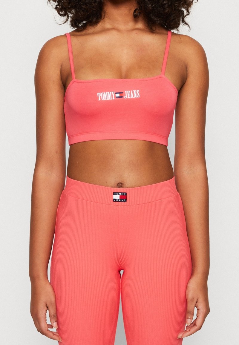 Koralfarbenes Bralette mit dünnen Trägern, das mit dem weißen "TOMMY JEANS"-Logo versehen ist. Rippstoff, dehnbares Material; kombiniert mit passenden koralfarbenen, gerippten Leggings.