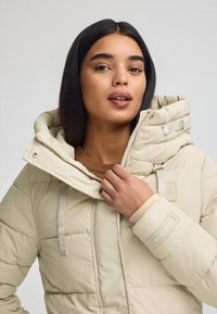 Beżowa kurtka puchowa z wysokim kołnierzem, zamkiem błyskawicznym z przodu i regulowanymi sznurkami. Posiada naszywkę z logo oraz ściągacze przy mankietach. Teksturalny, pikowany wzór.