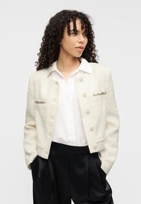 Λευκό υφαντό σακάκι με κουμπιά και μεταλλικές λεπτομέρειες, φορεμένο πάνω από πουκάμισο με γιακά, συνδυασμένο με μαύρο παντελόνι ψηλόμεσο.