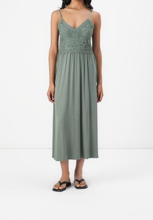 Femme portant une robe midi sans manches vert sauge avec un détail en dentelle sur le corsage et de fines bretelles, associée à des sandales noires.