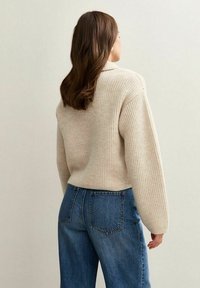 Femme avec de longs cheveux bruns portant un pull en ribbed beige et un jean en denim bleu, de dos contre un fond clair uni.