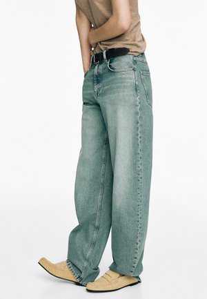 Personne portant un jean ample bleu clair avec une ceinture noire, une chemise beige et des mocassins couleur camel, debout devant un fond blanc uni.