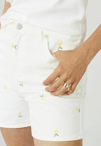 Des shorts en denim blanc ornés de broderies de citrons jaunes éclatants avec des feuilles vertes. Le style comprend une taille classique et des poches avant.
