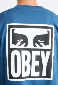 Blå bomull T-skjorte med et stort svart-hvitt grafisk design med hender og ordet "OBEY" i fet skrift på baksiden.