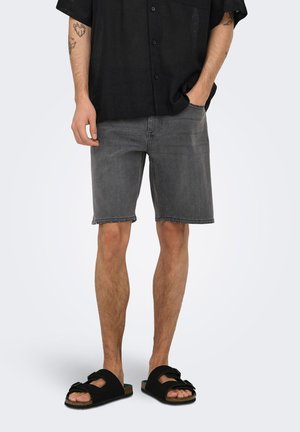 Homme portant une chemise noire boutonnée, un short en denim gris et des sandales noires à brides, debout avec une main dans la poche et des tatouages visibles sur le bras.