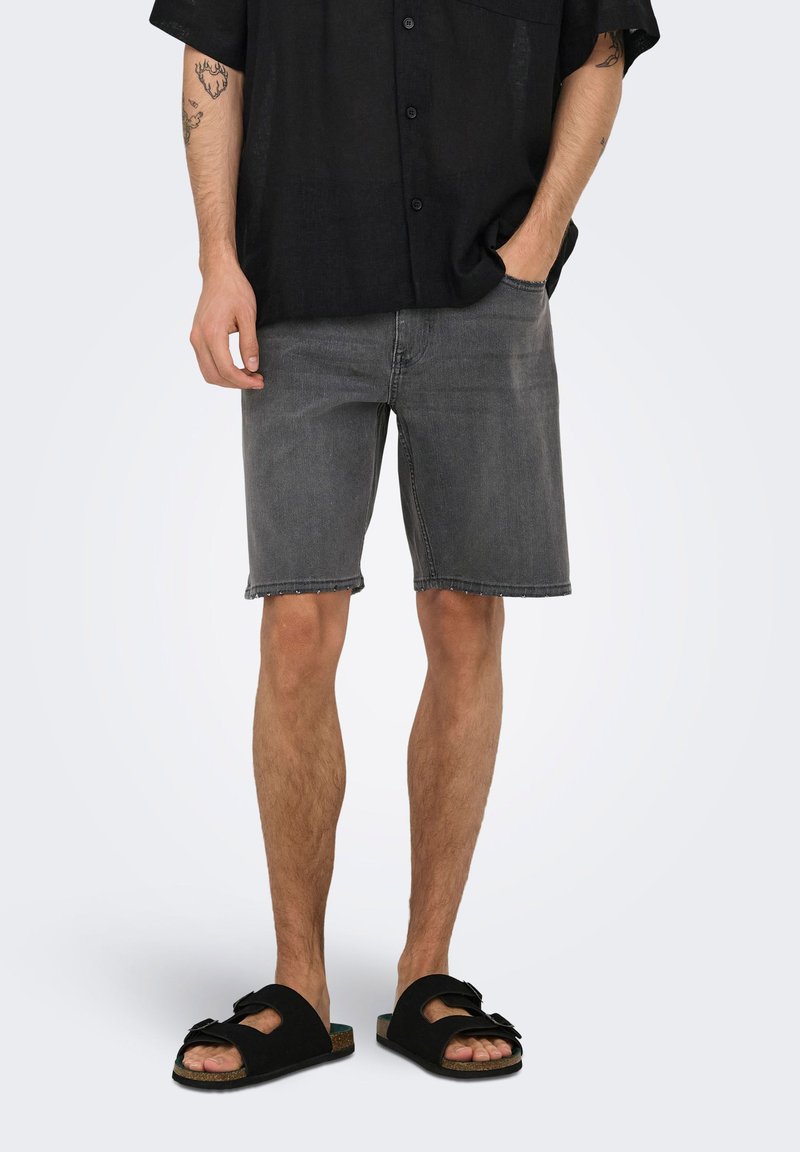 Homme portant une chemise noire boutonnée, un short en denim gris et des sandales noires à brides, debout avec une main dans la poche et des tatouages visibles sur le bras.