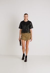 Top curto de manga curta em couro sintético preto, saia mini riscada em tons de bege, botas de tornozelo pretas com meias, segurando uma pequena carteira. Fundo minimalista.