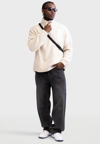 Κρεμ φλις πόλο με τσέπη στην πλάτη με φερμουάρ, σε συνδυασμό με σκούρα φαρδιά τζιν και λευκά αθλητικά παπούτσια. Το μοντέλο φορά μαύρα γυαλιά ηλίου.