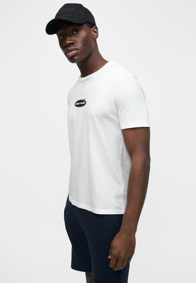 Bílé bavlněné tričko s černým vyšívaným logem "VOLTAIRE" na hrudi. Model nosí námořnicky modré šortky a pózuje před jednoduchým pozadím.