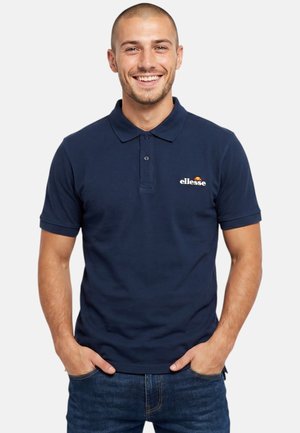 Homme souriant les mains dans les poches, portant un polo ellesse bleu marine et un jean bleu sur un fond clair uni.