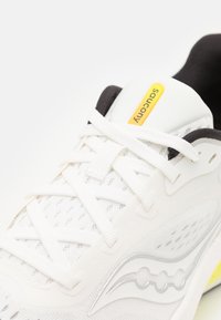 Scarpa atletica bianca con tomaia in mesh texture, dettagli neri, logo giallo e elementi di design grigio sottili. Presenta un tradizionale sistema di allacciatura.