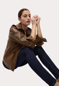 Chaqueta de cuero marrón con un acabado texturizado, que presenta un cuello doblado, puños abotonados y bolsillos de parche, combinada con unos jeans de mezclilla oscuros.