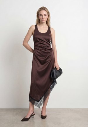 Femme debout portant une robe en satin marron foncé sans manches avec un ourlet en dentelle noire, tenant une pochette noire et portant des talons slingback marron.