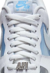 Witte en lichtblauwe Nike Air Force 1 sneaker in close-up met witte veters en een met juwelen versierde "AF1" veterclip op de tong.