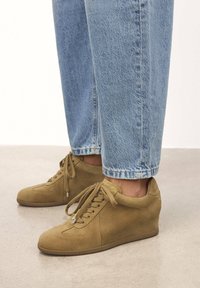 Sneakers a zeppa in suede marrone chiaro con punta arrotondata, lacci frontali e sottili dettagli di design. Abbinati a jeans in denim azzurri con risvolto.