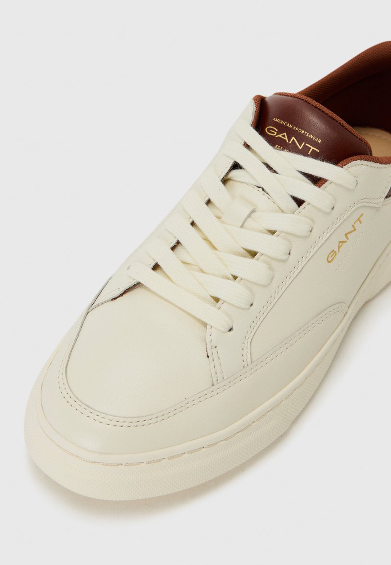 Zapatilla blanca de piel con parte superior texturizada, punta redonda y cordones en crema. Detalle en piel marrón en el talón con el logo dorado "GANT".