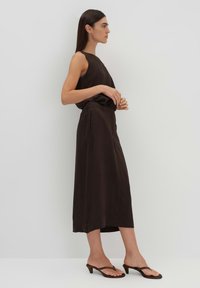 Robe sans manches marron à coupe décontractée, dotée d'une taille ceinturée et de poches latérales. Le tissu a une texture lisse et tombe en dessous du genou.