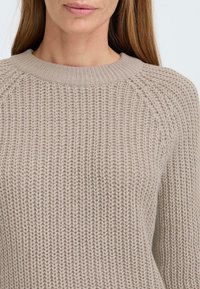 Beige strikket genser med ribbet rund hals og strukturerte, løse maskemønstre. Strukturert ermer med raglan-design.