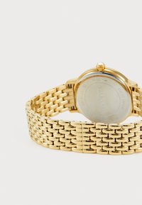 Gold-tone wristwatch with a textured metal bracelet, round case, and a single accent crystal on the crown, showcasing a polished finish.