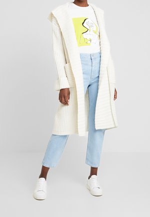 Henkilö, joka pukeutuneena pitkään valkoiseen neulottuun neuletakkiin, valkoiseen graafiseen t-paitaan, jossa on keltainen neliö, vaalean sinisiin farkkuihin ja valkoisiin tennareihin, seisoo harmaalla lattialla.