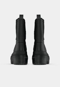 Cizme negre până la gleznă, cu un superior texturat, caracterizându-se printr-o talpă groasă din cauciuc, mânere pe spate și un design simplu fără branding vizibil.