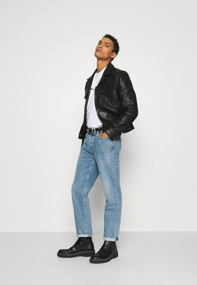 Jachetă din piele neagră peste un tricou alb, blugi din denim albastru deschis cu manșete răsucite și bocanci negri la glezne, cu un detaliu din lanț pe curea.