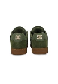 Par av gröna mockaskor med bruna sulor, sedda bakifrån, med en vit DC-logga på hälflikarna.