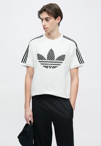 Άσπρο βαμβακερό μπλουζάκι με μαύρο διακοσμημένο λογότυπο Adidas, με μαύρες ριγέ λεπτομέρειες στα κοντά μανίκια, σε συνδυασμό με μαύρο παντελόνι.