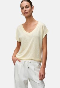 Jasnożółta, krótko rękawowa koszulka typu V-neck, wykonana z miękkiego, teksturowanego materiału, zestawiona z białymi spodniami ze sznurkiem w pasie, ręce w kieszeniach.