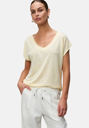 Light yellow, short-sleeve V-neck t-shirt made of soft, textured fabric, paired with white drawstring pants, hands in pockets.