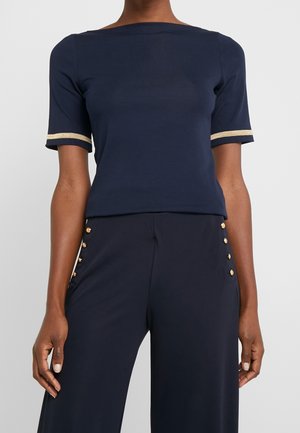 Femme portant un haut bleu marine à manches courtes avec un liseré doré sur les manches et un pantalon bleu marine avec des boutons dorés sur les côtés.