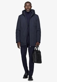 Marineblå polstret jakke med hætte, båret over en marineblå turtleneck og ternede bukser; holder en sort håndtaske, elegante sko.