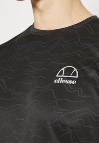 Černé sportovní tričko s texturovaným vlnitým vzorem v tmavším odstínu, s bílým logem "ellesse" v horním levém rohu.