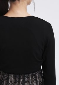 Mustat pitkät hihat -paita, jossa on pyöreä kaula-aukko, yhdistettynä läpinäkyvään mustaan pitsihameeseen. Pitsissä on kukkakuosia ja kerroksellisia yksityiskohtia.