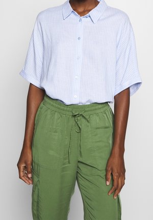 Person iført en lyseblå kortærmet skjorte med tynde hvide striber og grønne snøre bukser med lommer.