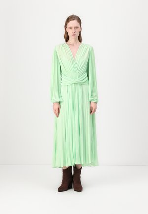 Faltenrock, langärmliges Kleid in Hellgrün mit tiefem V-Ausschnitt und verdrehtem Taillendetail; kombiniert mit braunen, hochhackigen Stiefeletten.