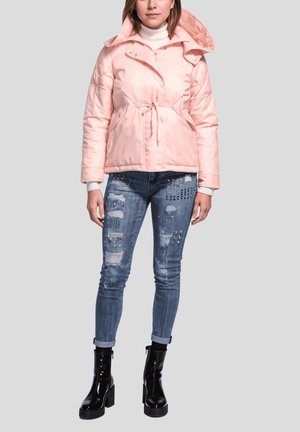 Mujer llevando una chaqueta acolchada rosa claro con cintura ajustable, vaqueros azules desgastados con adornos y botas negras de charol con tacón.