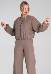 Brązowa kurtka zapinana na zamek z okrągłym dekoltem i elastycznym paskiem w talii, w zestawie z dopasowanymi spodniami typu wide-leg. Miękki, teksturowany materiał, minimalne detale.