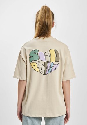 T-shirt beige oversize con un design di cuore colorato e la scritta "Still Ballin'" in diverse tonalità pastello sul retro.