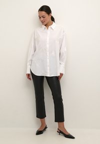Camicia bianca con ricami floreali delicati, vestibilità comoda, abbinata a pantaloni cropped in finta pelle nera e scarpe nere a punta.