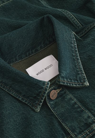 Colletto di giacca di jeans verde scuro con etichetta di tessuto bianca visibile con la scritta "WOOD WOOD" e un bottone metallico inciso con "W.W."