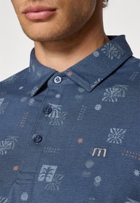 Navy polo-skjorte med et mønsterdesign med botaniske motiver, knapplukking på kragen, og sølvlogo på venstre side.