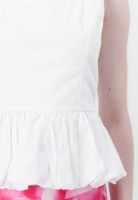 Hvit peplum-top laget av glatt stoff, med en samlet kant, og sammen med et rosa mønstrete skjørt. Enkel, elegant design.