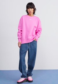 Sweatshirt rose avec un logo blanc, associé à un jean en denim ample. Le modèle porte des chaussures roses et noires, se tenant sur une surface bleue.
