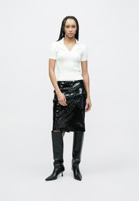 Weißes, geripptes Strickoberteil mit V-Ausschnitt, kombiniert mit einem schwarzen Bleistiftrock aus Pailletten. Schwarze kniehohe Stiefel mit Absatz vervollständigen den Look.