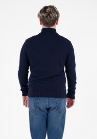 Dunkelblaues gestricktes Sweatshirt mit hohem Kragen sowie gerippten Säumen und Bündchen, kombiniert mit hellblauen Jeans. Von hinten betrachtet.