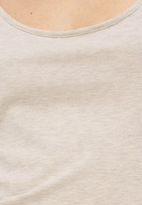 Κοντινό πλάνο υφάσματος ανοιχτού μπεζ με λεπτή υφή και απλό στρογγυλό ντεκολτέ σε φόντο χρώματος επιδερμίδας.