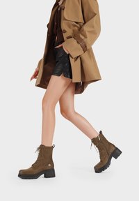 Stivaletti in suede marrone con punta tonda, chiusura con lacci frontale, tacco spesso nero e dettaglio con zip laterale. Esterno testurizzato.
