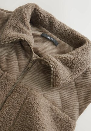 Brūna vilnas jaka ar quilted dizainu, ar rāvējslēdzēju un mīkstu tekstūru. Iedvesmots ar apkakli un melnu birku ar uzrakstu "NEXT."